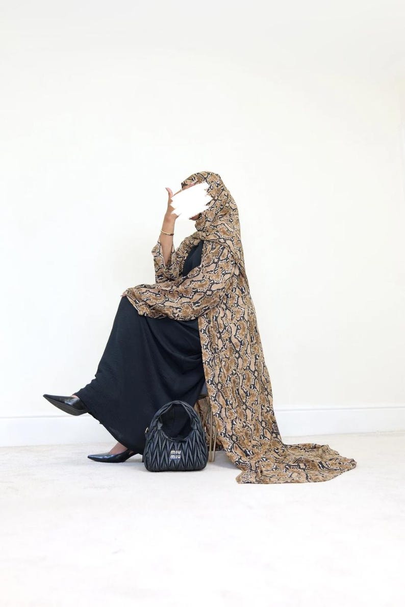 May include: A person wearing a black dress and a long, snake-print patterned coat with a matching head covering. A black handbag with the brand name is on the floor. The person is wearing black shoes.