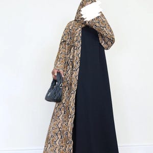May include: A black dress with a long, open snakeskin-print overcoat. The overcoat features brown and tan tones. A black handbag and black heels complete the outfit. The model is wearing a matching snakeskin print head covering.