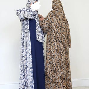 Könnte beinhalten: Zwei bodenlange Abayas. Eine ist marineblau mit einer weißen und blauen Schlangenhaut-Außenschicht. Die andere ist braun und hellbraun mit Schlangenhaut-Muster. Beide haben passende Kopfbedeckungen. Die Abayas werden von zwei Personen getragen.
