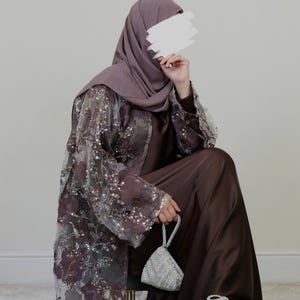 May include: A brown satin dress and matching hijab are worn with a sheer, floral-embroidered overcoat. A small, silver beaded handbag and matching flats complete the outfit. The ensemble is elegant and modest.