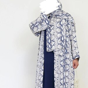 May include: A long, flowing outfit with a snakeskin print in white, grey, and navy blue. The ensemble includes a matching head covering and a long, dark blue dress. A small, white beaded bag is held in the hand.