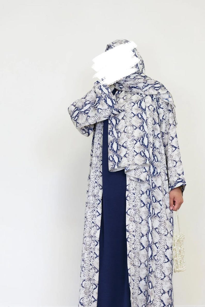May include: A long, flowing outfit with a snakeskin print in white, grey, and navy blue. The ensemble includes a matching head covering and a long, dark blue dress. A small, white beaded bag is held in the hand.