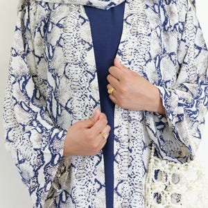 May include: A snakeskin-patterned kimono in white, blue, and tan, with a matching headscarf. The open front reveals a navy blue top. A white beaded purse hangs on the side. The garment is a stylish addition to any wardrobe.