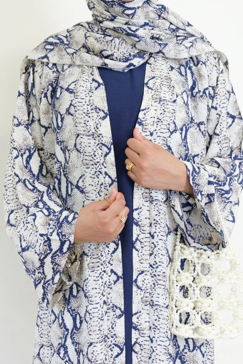 May include: A snakeskin-patterned kimono in white, blue, and tan, with a matching headscarf. The open front reveals a navy blue top. A white beaded purse hangs on the side. The garment is a stylish addition to any wardrobe.