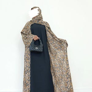May include: A dark navy blue abaya with a snakeskin print cape and matching head covering. The cape and head covering feature a brown and tan snakeskin pattern. A black handbag is held in the hand.