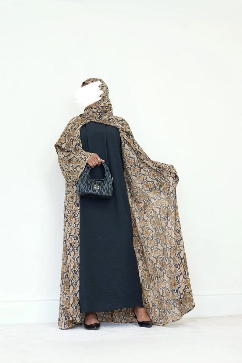May include: A dark navy blue abaya with a snakeskin print cape and matching head covering. The cape and head covering feature a brown and tan snakeskin pattern. A black handbag is held in the hand.