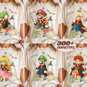 Op de afbeelding: Een crèmekleurig t-shirt met een grafische print van diverse Nintendo-personages in tovenaarsgewaden en sjaals, die toverstokken vasthouden en op stapels boeken zitten. Het ontwerp bevat zwevende brillen, een gouden snaai en een heksenhoed.