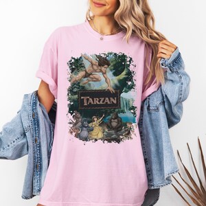 May include: A light pink t-shirt featuring a graphic print of the character Tarzan leaping above a rectangular sign that reads Tarzan, with Jane and two gorillas positioned below in a jungle setting with a waterfall in the background.