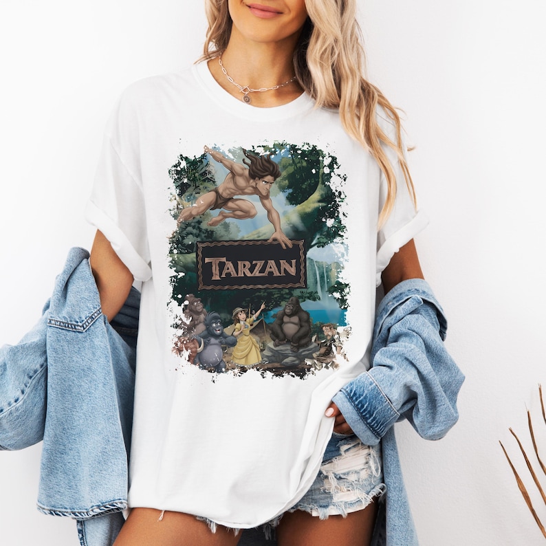 May include: A white t-shirt featuring a graphic print of the Disney movie Tarzan. The design shows Tarzan leaping above a rectangular frame containing the text TARZAN, with characters Jane, Terk, and Kerchak positioned in a jungle setting.