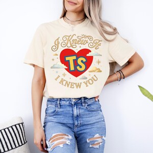 Puede incluir: Una camiseta de color crema con un dise&ntilde;o gr&aacute;fico que presenta un coraz&oacute;n rojo, las letras amarillas TS y el texto I KNEW IT I KNEW YOU en letra dorada. El dise&ntilde;o incluye peque&ntilde;as nubes y estrellas alrededor del coraz&oacute;n central.