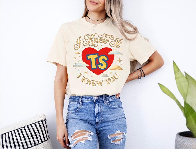 Puede incluir: Una camiseta de color crema con un dise&ntilde;o gr&aacute;fico que presenta un coraz&oacute;n rojo, las letras amarillas TS y el texto I KNEW IT I KNEW YOU en letra dorada. El dise&ntilde;o incluye peque&ntilde;as nubes y estrellas alrededor del coraz&oacute;n central.