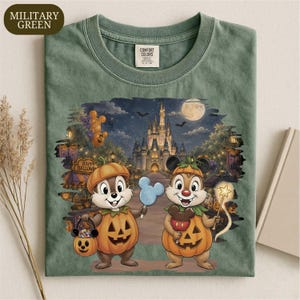 May include: A military green t-shirt featuring a graphic of two chipmunks dressed in pumpkin costumes. One holds a pumpkin-shaped candy bucket and a blue Mickey-shaped treat, while the other holds a star wand. A castle is in the background.