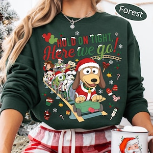 Può includere: Una felpa girocollo verde bosco con una stampa grafica di vari personaggi dei cartoni animati su una montagna russa. Il testo recita "HOLD ON TIGHT, Here we go!" e "Noel". Il design include elementi natalizi come fiocchi di neve, campanelle e bastoncini.