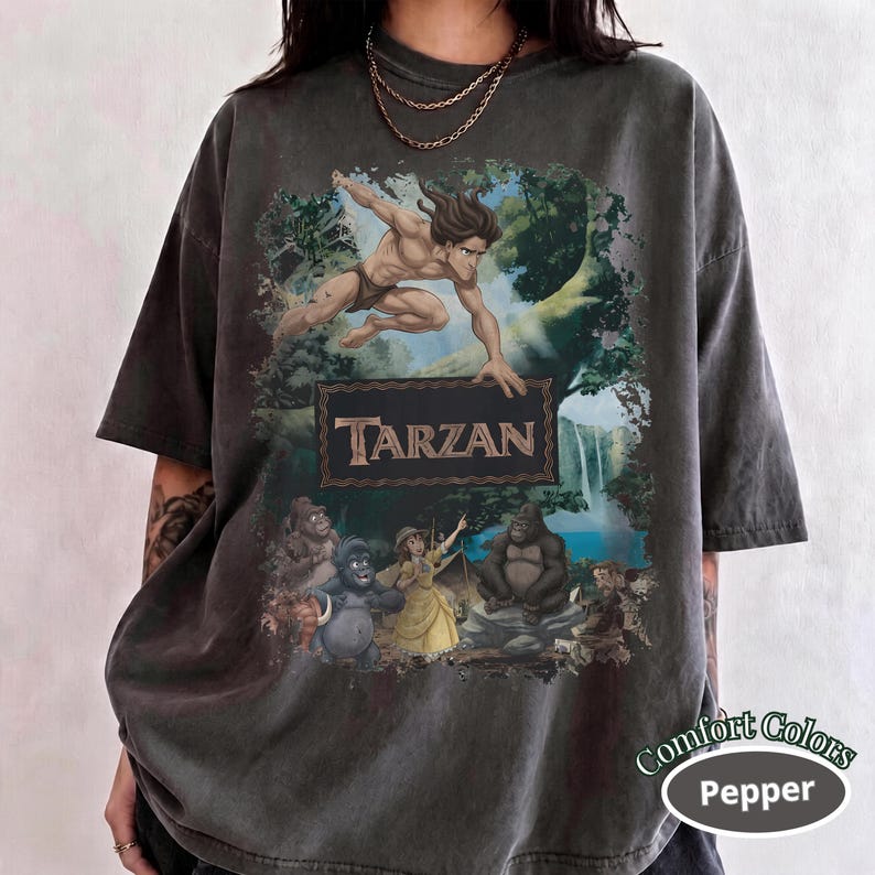 May include: A charcoal gray short-sleeve t-shirt featuring a graphic print of Tarzan leaping above a rectangular frame containing the text TARZAN, with illustrations of Jane and gorillas in a jungle setting below the text, all in a vintage style.