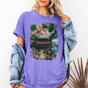 May include: A purple short-sleeve t-shirt featuring a graphic print of the movie Tarzan. The design includes Tarzan leaping above a rectangular sign with the text Tarzan, with Jane, gorillas, and jungle scenery depicted below the title.