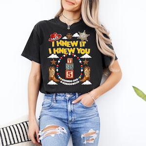 May include: A black short-sleeve t-shirt featuring a graphic print with the text I KNEW IT I KNEW YOU, a red cowboy hat, a sheriff badge, two brown cowboy boots, and three stacked blocks labelled T, S, and 5, all surrounded by a colourful beaded border.