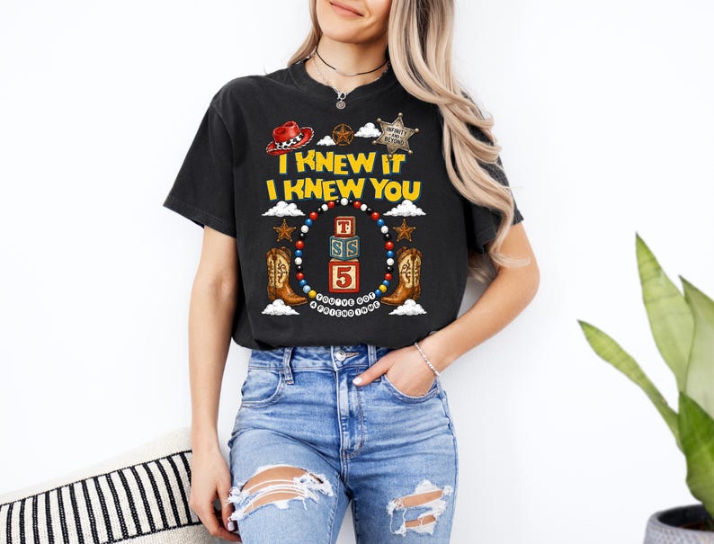 May include: A black short-sleeve t-shirt featuring a graphic print with the text I KNEW IT I KNEW YOU, a red cowboy hat, a sheriff badge, two brown cowboy boots, and three stacked blocks labelled T, S, and 5, all surrounded by a colourful beaded border.