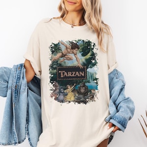 May include: A cream-colored short-sleeve t-shirt featuring a graphic print of the character Tarzan leaping above a jungle scene with gorillas and Jane. The text Tarzan is centered in a dark rectangular frame within the colorful illustration.