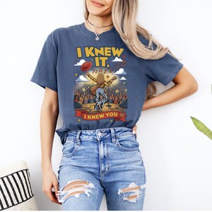 Op de afbeelding: Een blauw t-shirt met korte mouwen en een afbeelding van een artiest op een podium met een gitaar, een rode hoed en muzieknoten. De tekst luidt I KNEW IT, I KNEW YOU TS. Het ontwerp bevat een menigte, sterren en wolken in geel, rood en wit.