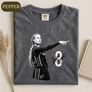 May include: A charcoal gray crew neck t-shirt featuring a black and white graphic of a basketball player pointing forward. The design includes the number 8 in white with a red outline and a black signature. The shirt has a Comfort Colors brand label.