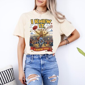 Op de afbeelding: Een cr&egrave;mekleurig T-shirt met korte mouwen en een afbeelding van een muzikant op het podium met een gitaar, een zwevende rode hoed en muzieknoten. De tekst luidt I KNEW IT, en I KNEW YOU TS in gele en rode letters tegen een concertachtergrond.