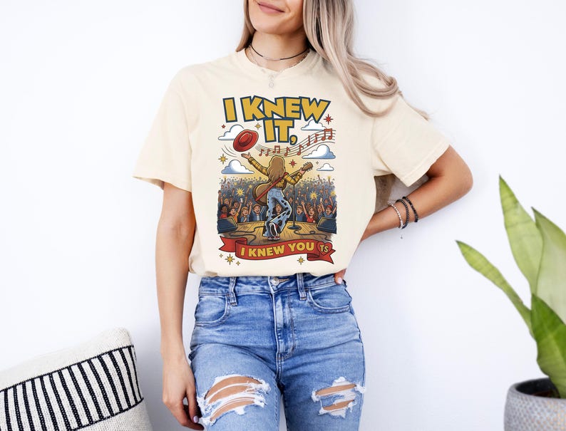 Op de afbeelding: Een cr&egrave;mekleurig T-shirt met korte mouwen en een afbeelding van een muzikant op het podium met een gitaar, een zwevende rode hoed en muzieknoten. De tekst luidt I KNEW IT, en I KNEW YOU TS in gele en rode letters tegen een concertachtergrond.