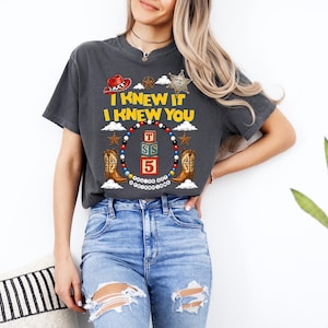 May include: A charcoal grey short-sleeve t-shirt featuring a graphic print with the text I KNEW IT I KNEW YOU, a red cowboy hat, a sheriff star, cowboy boots, and a circular frame of colourful beads surrounding stacked alphabet blocks.