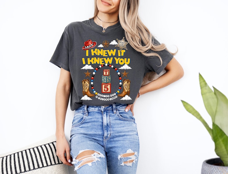 May include: A charcoal grey short-sleeve t-shirt featuring a graphic print with the text I KNEW IT I KNEW YOU, a red cowboy hat, a sheriff star, cowboy boots, and a circular frame of colourful beads surrounding stacked alphabet blocks.