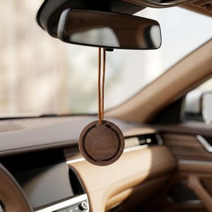 May include: A circular dark brown wooden car air freshener hanging from a rearview mirror by a thin tan leather cord. The wooden disc features a smooth finish with a circular cutout detail along the inner edge, suspended in the center of the vehicle.