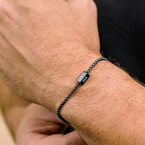May include: A black chain link bracelet featuring a small rectangular charm engraved with the names DANIEL and AVA in white lettering. The bracelet consists of a series of interconnected dark metal links that form a continuous, flexible band.