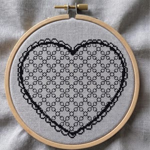 May include: A circular wooden embroidery hoop containing a piece of light gray fabric. The fabric features a black cross-stitch pattern of a heart shape filled with a repeating geometric grid design and a decorative scalloped lace-like border.