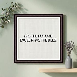 May include: Framed cross-stitch art with the text "AI IS THE FUTURE, EXCEL PAYS THE BILLS" in black. The artwork is in a dark brown frame with a white mat. A small vase with dried flowers sits on a wooden shelf below.