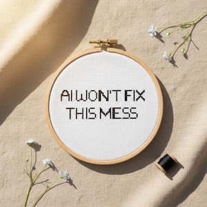 May include: A cross-stitch embroidery hoop with the text "AI WON'T FIX THIS MESS" in black. The white fabric is stretched within a wooden hoop. A black thread spool and sprigs of blue flowers are nearby, set against a beige fabric background.