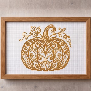 May include: A cross-stitch embroidery of a pumpkin featuring intricate golden-yellow floral and scrollwork patterns on white Aida cloth, encased in a rectangular light-toned wooden frame with a visible grain texture and a clean, simple border.