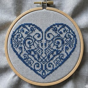 May include: A cross-stitch embroidery in a wooden hoop. The design features a detailed blue heart with intricate patterns on a light gray fabric. The wooden hoop is secured with a brass screw.