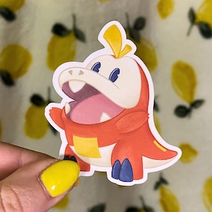 Puede incluir: Una colorida pegatina con un personaje de dibujos animados con forma de cocodrilo y la boca abierta. El personaje es principalmente naranja con detalles blancos, amarillos y azules. La pegatina se sostiene sobre un fondo con un estampado de limones.