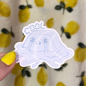 Puede incluir: Una pegatina azul claro con un personaje de dibujos animados con una cabeza en forma de cubo, ojos en forma de estrella y lágrimas. La pegatina tiene una base fluida y líquida y está rematada con tres estrellas. El fondo es una tela con limones amarillos.