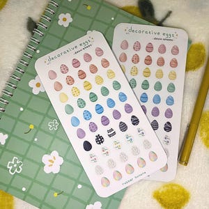 May include: Two sticker sheets featuring decorative Easter egg designs in a rainbow of colours and patterns. The sheets are labelled "decorative eggs deco sheet" and include the text "rubber banana". A green notebook and yellow pen are also visible.