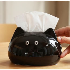 Op de afbeelding: Een zwarte keramische tissuebox in de vorm van een kattenkop, met witte ogen en oren. Witte tissues steken uit de bovenkant. De tissuebox staat op een houten oppervlak.
