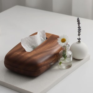 May include: A dark wooden tissue box with a curved top, dispensing white tissues. A small glass vase with a daisy and a white spherical vase with lavender sprig sit beside it on a white surface.