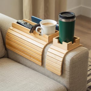 Puede incluir: Un juego de dos bandejas de bambú para el brazo del sofá. Una bandeja contiene un mando a distancia, un teléfono y una taza blanca con café. La otra contiene un termo de viaje verde. Las bandejas están diseñadas para encajar sobre el brazo de un sofá.