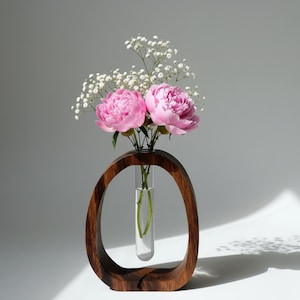 Könnte beinhalten: Eine dekorative Holzvase mit ovalem Rahmen hält ein Glasröhrchen mit Blumen. Zwei rosa Pfingstrosen und weißes Schleierkraut sind in der Vase angeordnet. Die Vase ist aus dunkelbraunem Holz gefertigt und ein stilvolles Wohnaccessoire.