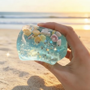 May include: A clear, blue resin paperweight filled with seashells and star-shaped glitter. The paperweight is held in a hand, with a beach and ocean in the background. The seashells are various colours and sizes.