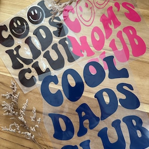 Könnte beinhalten: Drei Aufbügel-Vinyltransfers mit den Schriftzügen "COOL KIDS CLUB" in Schwarz, "COOL MOM'S CLUB" in Pink und "COOL DADS CLUB" in Marineblau. Jeder Schriftzug hat ein Smiley-Gesicht. Die Transfers befinden sich auf einer Holzoberfläche.