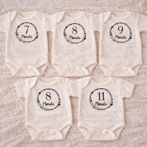 Könnte beinhalten: Fünf cremefarbene Baby-Bodys mit schwarzen Kreisdesigns, die die Monate 7, 8, 9 und 11 anzeigen. Jedes Design enthält das Wort "Monate". Die Bodys sind auf einer strukturierten, cremefarbenen Oberfläche angeordnet.
