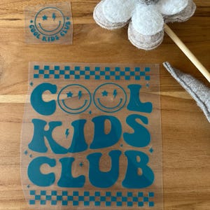 Könnte beinhalten: Ein blaugrünes Aufbügel-Abziehbild mit dem Text COOL KIDS CLUB in einer Retro-Schriftart mit Smileys und Blitzen, begleitet von einem kleineren passenden Abziehbild und einer weiß-beigen Filzblume an einem Holzstab.