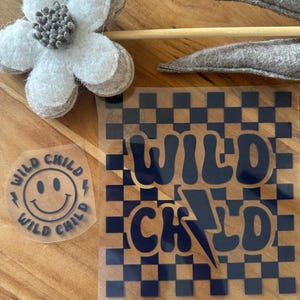 Könnte beinhalten: Zwei Klebeaufkleber mit dem Text Wild Child. Einer ist ein kleiner runder Aufkleber mit einem Smiley-Gesicht und Blitzen. Der andere ist ein größerer quadratischer Aufkleber mit einem dunkelblauen Schachbrettmuster und dem Text Wild Child mit einem Blitz.