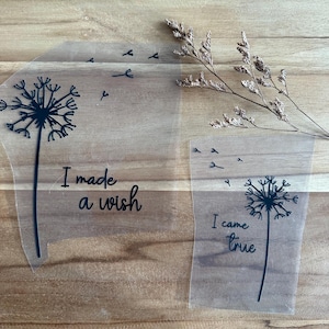 Könnte beinhalten: Zwei transparente Aufkleber mit schwarzen Pusteblumen-Designs und den Sätzen "I made a wish" und "I came true". Sie liegen auf einer Holzoberfläche mit getrockneten floralen Akzenten.