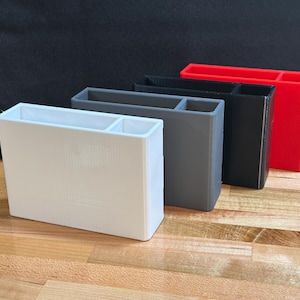 May include: Four rectangular desk organizers in white, gray, black, and red. Each organizer has two compartments. The organizers are made of a solid material and are arranged on a wooden surface.