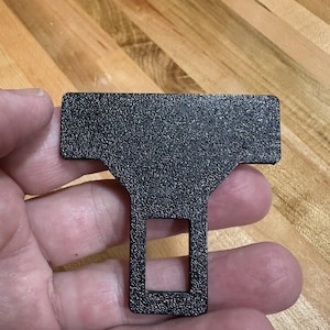 May include: A black seat belt buckle insert with a textured, matte finish. The object features a wide, rectangular top section that tapers down into a narrower, vertical stem containing a rectangular cutout opening in the center of the lower portion.